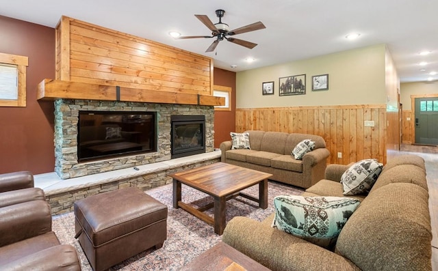 living room with a ceiling fan, a wainscoted wall, wood walls, a stone fireplace, and recessed lighting