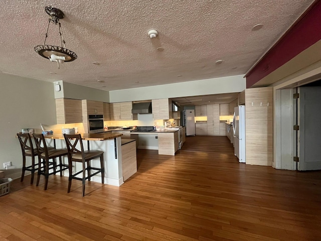 kitchen with white refrigerator, wood-type flooring, oven, wall chimney range hood, and a breakfast bar