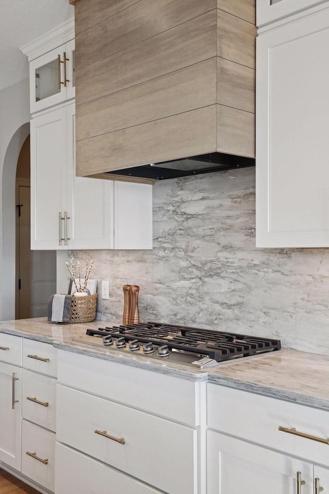 kitchen featuring light stone countertops, premium range hood, stainless steel gas cooktop, backsplash, and white cabinets