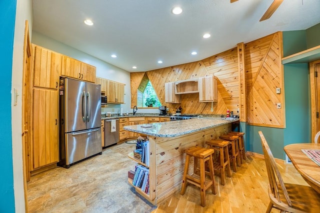 kitchen featuring appliances with stainless steel finishes, kitchen peninsula, wood walls, light stone countertops, and ceiling fan