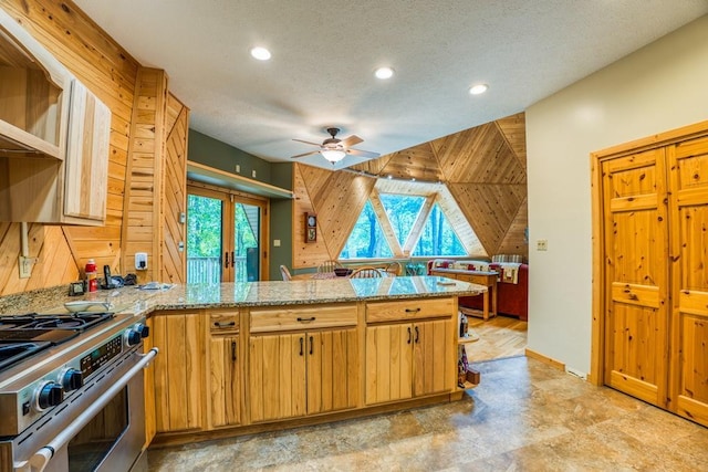 kitchen featuring light stone counters, stainless steel stove, kitchen peninsula, wood walls, and ceiling fan