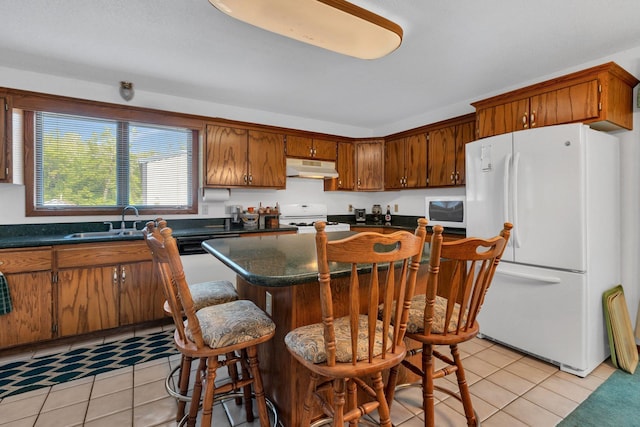kitchen featuring under cabinet range hood, light tile patterned floors, white appliances, a sink, and dark countertops