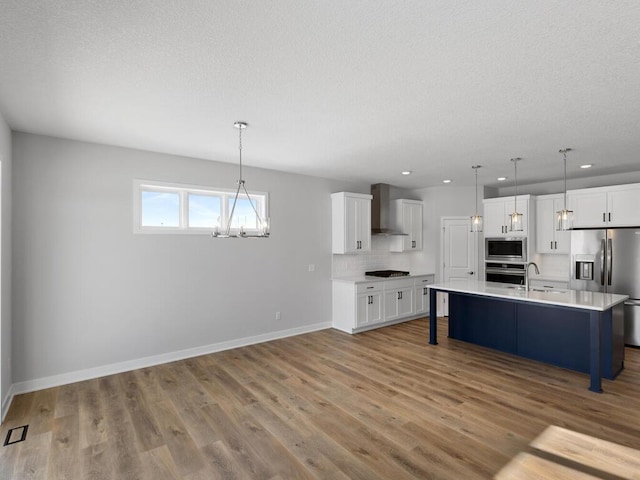 kitchen featuring wall chimney exhaust hood, decorative light fixtures, appliances with stainless steel finishes, an island with sink, and backsplash