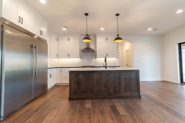 kitchen with white cabinetry, dark hardwood / wood-style floors, stainless steel built in fridge, hanging light fixtures, and a kitchen island with sink