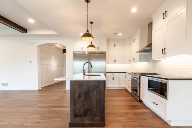 kitchen with wall chimney range hood, built in appliances, dark hardwood / wood-style flooring, pendant lighting, and an island with sink