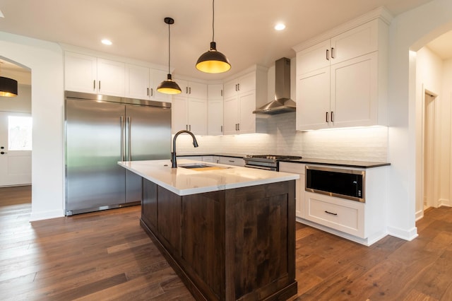 kitchen featuring built in appliances, a center island with sink, sink, dark wood-type flooring, and wall chimney range hood