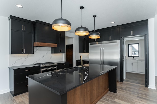 kitchen with appliances with stainless steel finishes, sink, dark stone countertops, hanging light fixtures, and a kitchen island with sink