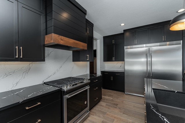 kitchen featuring appliances with stainless steel finishes, backsplash, custom range hood, dark stone counters, and light wood-type flooring