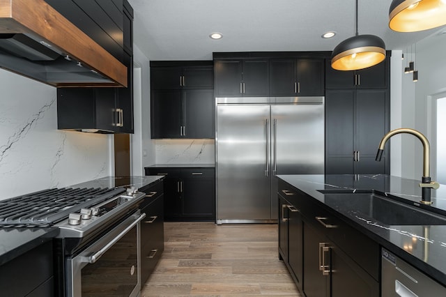 kitchen with appliances with stainless steel finishes, sink, dark stone counters, wall chimney exhaust hood, and light hardwood / wood-style flooring