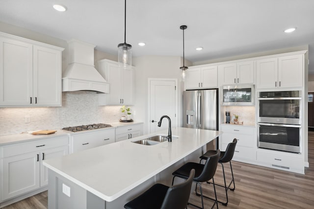 kitchen with custom range hood, hanging light fixtures, stainless steel appliances, a kitchen island with sink, and sink