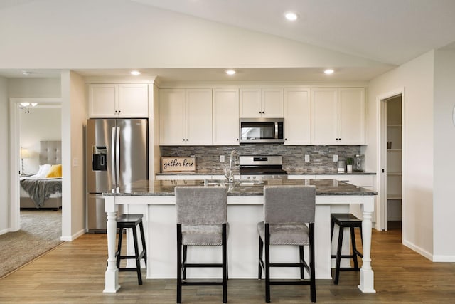 kitchen featuring appliances with stainless steel finishes, dark stone countertops, white cabinets, lofted ceiling, and a center island with sink