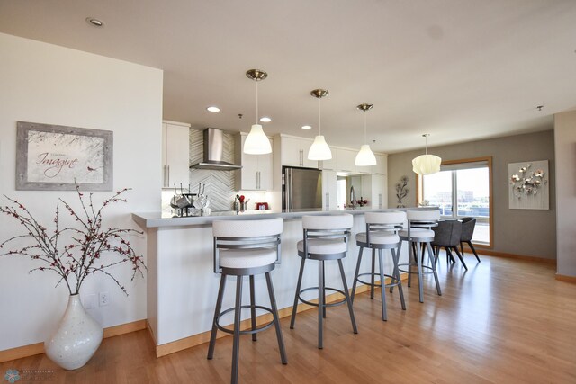 kitchen with white cabinets, hanging light fixtures, light hardwood / wood-style flooring, wall chimney range hood, and stainless steel refrigerator