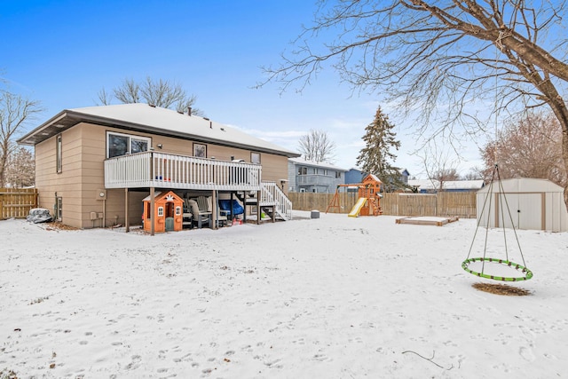 snow covered back of property with an outbuilding, a deck, a playground, a fenced backyard, and a storage unit