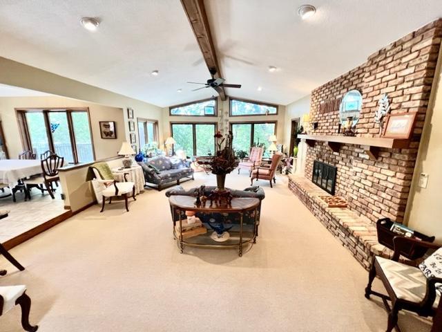 carpeted living room featuring a brick fireplace, vaulted ceiling with beams, and ceiling fan