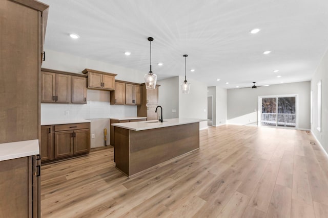 kitchen with light wood-type flooring, backsplash, a kitchen island with sink, ceiling fan, and hanging light fixtures