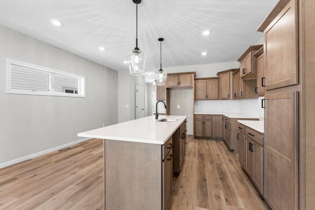 kitchen with light wood-type flooring, backsplash, a kitchen island with sink, sink, and hanging light fixtures