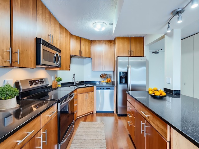 kitchen with brown cabinetry, dark stone counters, stainless steel appliances, light wood-type flooring, and a sink