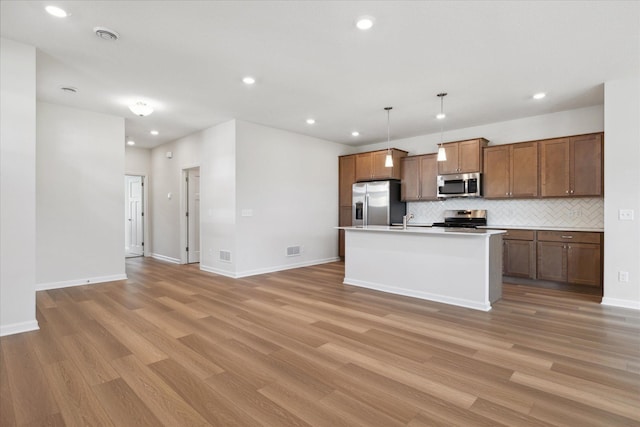 kitchen with appliances with stainless steel finishes, hanging light fixtures, tasteful backsplash, light hardwood / wood-style floors, and a center island with sink