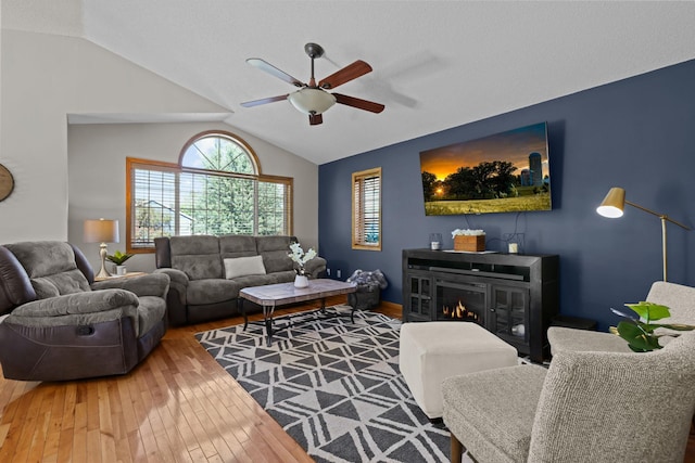 living area with a ceiling fan, a glass covered fireplace, vaulted ceiling, and hardwood / wood-style flooring