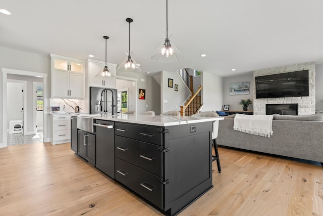 kitchen featuring stainless steel dishwasher, a kitchen island with sink, a stone fireplace, and white cabinetry