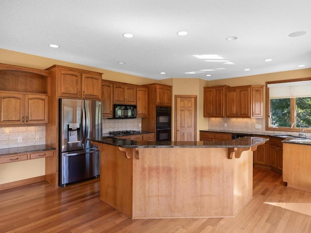 kitchen with dark stone counters, light wood-type flooring, a kitchen island, black appliances, and backsplash