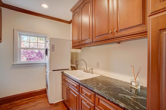 kitchen featuring sink, dark stone countertops, stacked washing maching and dryer, ornamental molding, and wood-type flooring