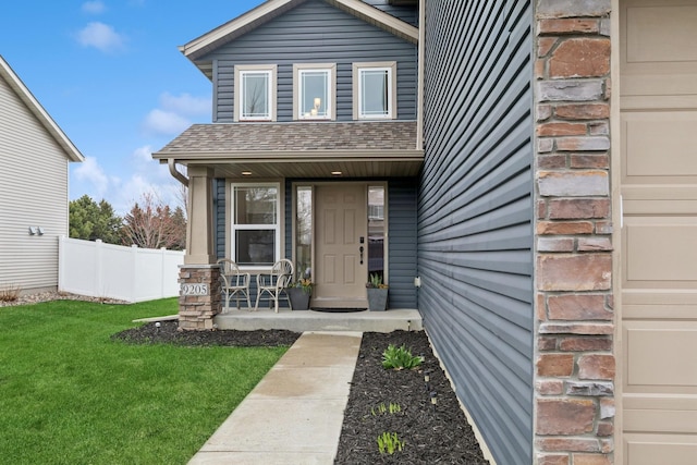 view of exterior entry featuring a lawn, a shingled roof, fence, and covered porch