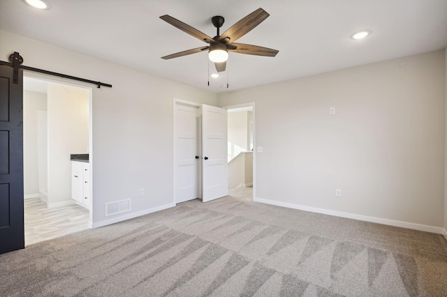 unfurnished bedroom featuring light carpet, a barn door, ensuite bath, and ceiling fan