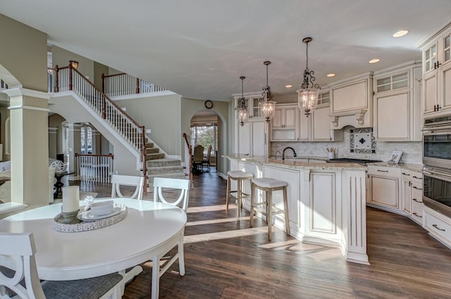 kitchen with dark wood-type flooring, backsplash, a center island with sink, pendant lighting, and light stone counters