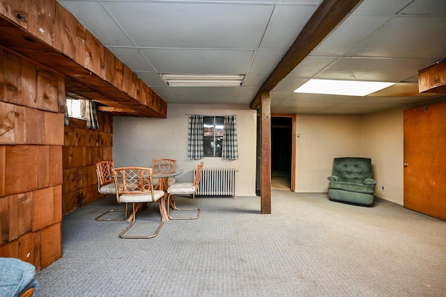 basement featuring a paneled ceiling, carpet, wooden walls, and radiator