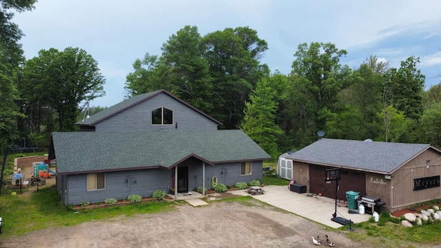 view of front of home with dirt driveway, a patio, a shingled roof, and an outbuilding