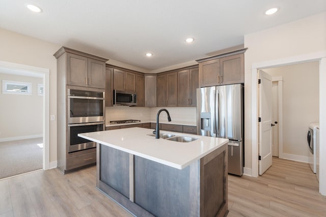 kitchen with light wood-type flooring, sink, an island with sink, washing machine and dryer, and stainless steel appliances