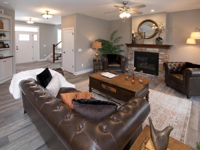living area featuring stairway, baseboards, ceiling fan, a glass covered fireplace, and wood finished floors