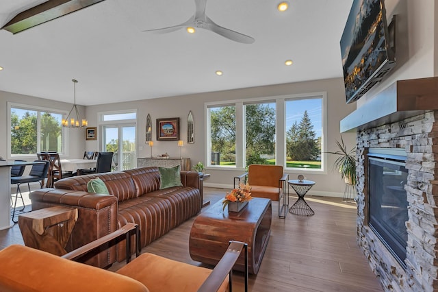 living room featuring hardwood / wood-style floors, ceiling fan with notable chandelier, beam ceiling, and a stone fireplace