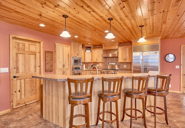 kitchen featuring pendant lighting, built in appliances, ventilation hood, a center island with sink, and light brown cabinets