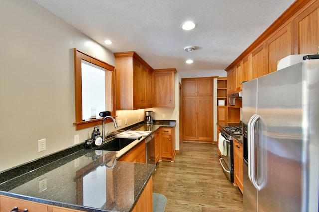 kitchen with a textured ceiling, light hardwood / wood-style floors, appliances with stainless steel finishes, dark stone countertops, and sink