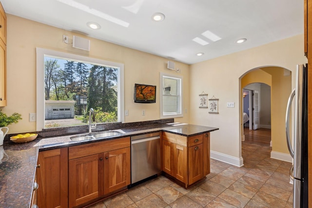 kitchen with stainless steel appliances, a sink, brown cabinetry, arched walkways, and dark stone counters