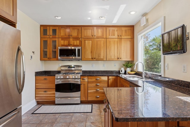 kitchen with appliances with stainless steel finishes, a sink, brown cabinets, dark stone countertops, and glass insert cabinets