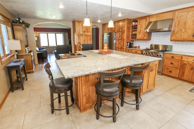 kitchen with stainless steel appliances, pendant lighting, a large island with sink, and wall chimney range hood