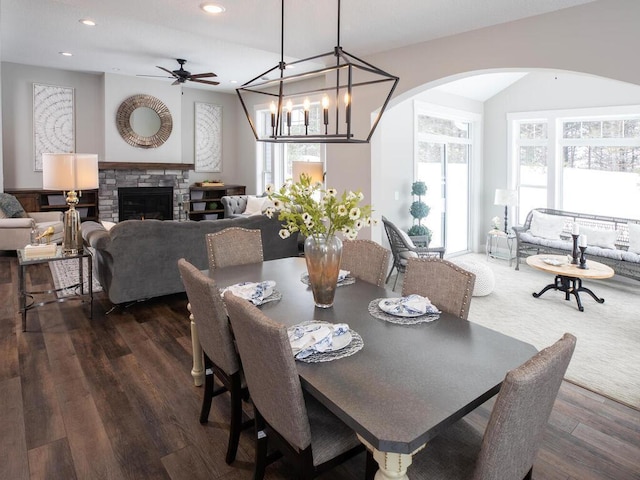 dining room with ceiling fan with notable chandelier, a fireplace, and dark wood-type flooring