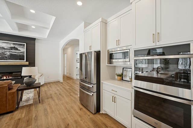 kitchen with stainless steel appliances, light wood-style floors, arched walkways, a glass covered fireplace, and white cabinetry