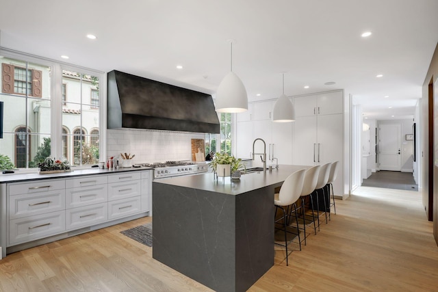 kitchen featuring hanging light fixtures, light hardwood / wood-style floors, white cabinets, a center island with sink, and wall chimney range hood