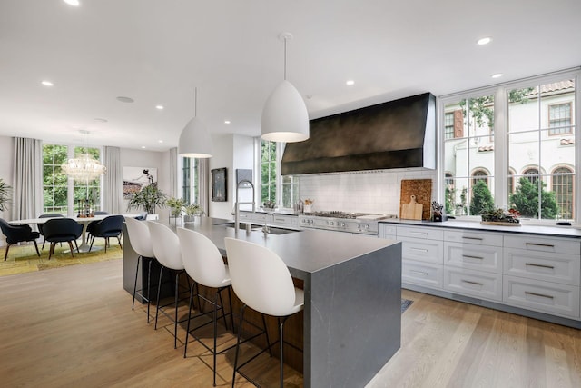 kitchen with pendant lighting, sink, a center island with sink, wall chimney range hood, and light wood-type flooring