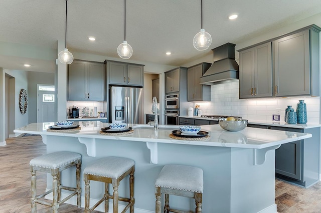 kitchen with appliances with stainless steel finishes, a large island with sink, pendant lighting, and custom exhaust hood