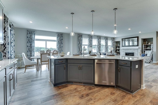 kitchen featuring sink, an island with sink, hanging light fixtures, a stone fireplace, and stainless steel dishwasher