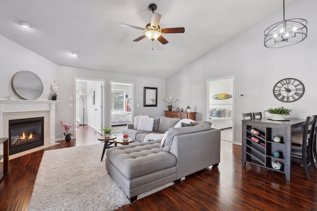 living room with high vaulted ceiling, ceiling fan with notable chandelier, a fireplace with flush hearth, wood finished floors, and baseboards