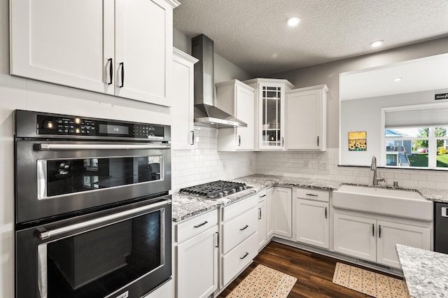 kitchen with white cabinets, wall chimney exhaust hood, light stone countertops, stainless steel appliances, and sink