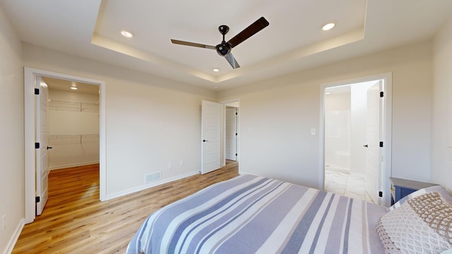 bedroom with ceiling fan, ensuite bathroom, a walk in closet, a raised ceiling, and light wood-type flooring