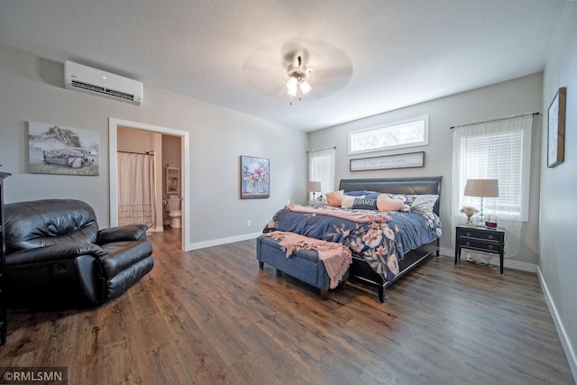 bedroom with a wall unit AC, ceiling fan, dark wood-type flooring, ensuite bathroom, and a textured ceiling