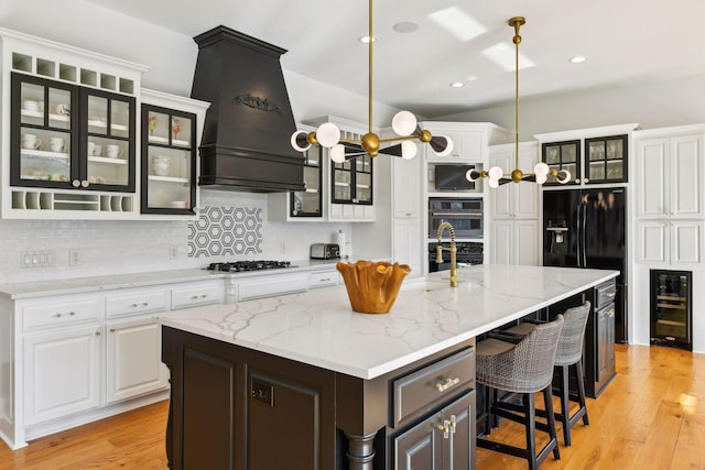 kitchen with an island with sink, white cabinets, custom range hood, wine cooler, and light hardwood / wood-style flooring